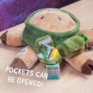 May include: A plush toy featuring a tan creature with a green hood and brown hooves. The toy has small pockets on its front that can be opened, secured with velcro. A small, clear, iridescent cube is placed on the front. Text reads: POCKETS CAN BE OPENED!