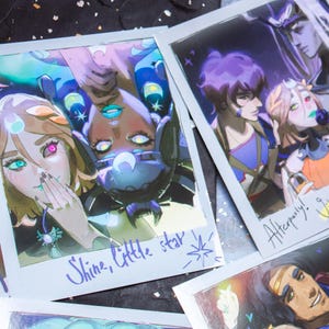 Può includere: Diverse stampe in stile polaroid con illustrazioni colorate di personaggi fantasy. Le stampe presentano opere d'arte dettagliate con colori vivaci, tra cui blu, viola e verde. Una stampa ha il testo "Shine, little star" e un'altra dice "Afterparty!"