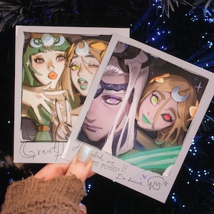 May include: Two polaroid-style prints featuring anime-style illustrations. One print shows three characters with green, yellow, and white accents. The other print shows two characters with purple, green, and yellow accents. Text includes "Great!" and "I'm doomed."