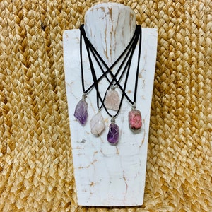 May include: Five necklaces with different colored crystal pendants. The pendants are wrapped in silver wire and hang from black cords. The necklaces are displayed on a white wooden mannequin.