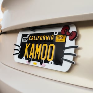 Puede incluir: Una placa de matrícula de California personalizada con el nombre "KAMOO" en letras amarillas. La placa es negra con un borde blanco y presenta un diseño de Hello Kitty, que incluye un lazo rojo y rasgos faciales. La placa está sujeta a un vehículo.