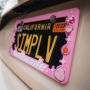 Puede incluir: Un marco rosa para matr&iacute;cula con el texto "SIMPLV" en amarillo y "California" en la parte superior. El marco est&aacute; decorado con personajes de dibujos animados y el texto "melcute". La matr&iacute;cula es negra con letras amarillas.
