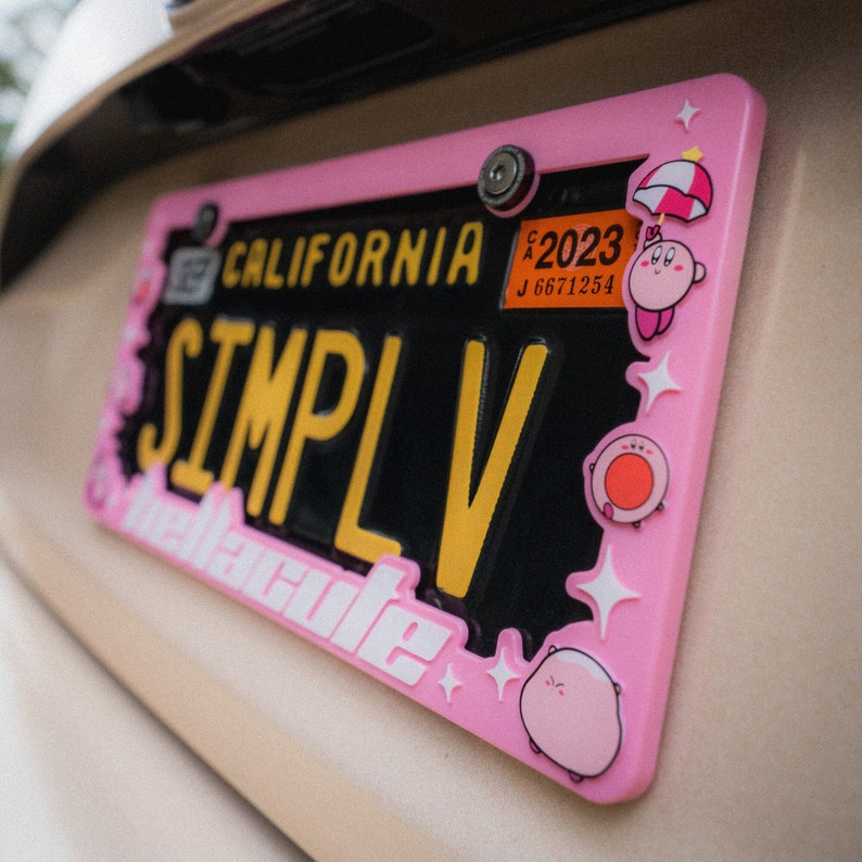 Puede incluir: Un marco rosa para matr&iacute;cula con el texto "SIMPLV" en amarillo y "California" en la parte superior. El marco est&aacute; decorado con personajes de dibujos animados y el texto "melcute". La matr&iacute;cula es negra con letras amarillas.
