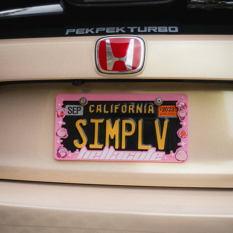 Puede incluir: Una placa de matr&iacute;cula rosa de California con la palabra "SIMPLV" en amarillo y el texto "bellacutie" en blanco. La placa tiene un borde rosa con dise&ntilde;os de personajes de dibujos animados. La placa est&aacute; montada en un coche beige.