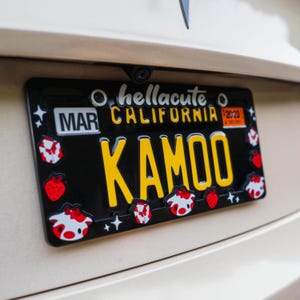 Może przedstawiać: Czarna tablica rejestracyjna z ż&oacute;łtymi literami z napisem "KAMOO" i "CALIFORNIA" z rokiem "2023". Tablica ma dekoracyjne truskawki, twarze kr&oacute;w i geometryczne kształty. Widoczny jest r&oacute;wnież tekst "hellacute".
