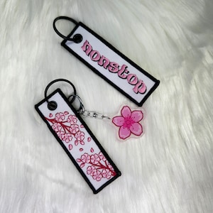 May include: Two keychains, one with a white background and black trim with the word "nonstop" embroidered in pink, and the other with a white background and black trim with a pink cherry blossom design.