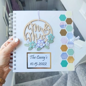 May include: White spiral bound journal with a wooden "Mr & Mrs" sign with a floral wreath and a gold hexagon pattern on the cover. The journal has the text "The Casey's 10.15.2022" on the front.