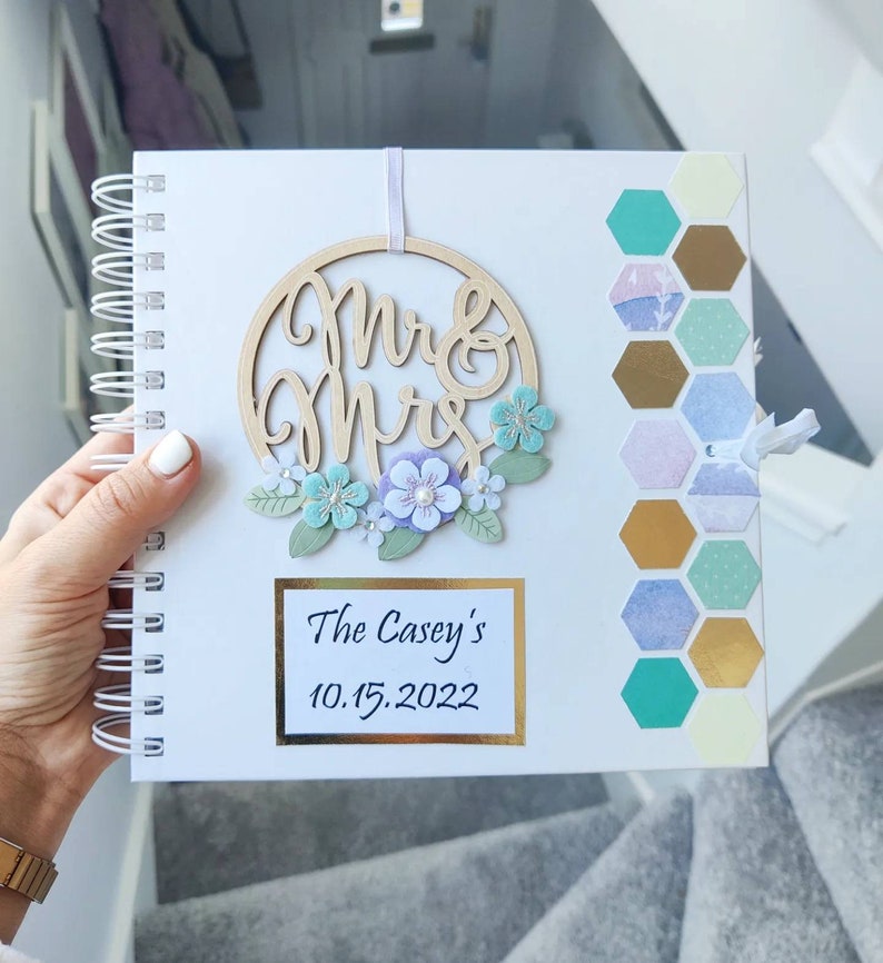 May include: White spiral bound journal with a wooden "Mr & Mrs" sign with a floral wreath and a gold hexagon pattern on the cover. The journal has the text "The Casey's 10.15.2022" on the front.