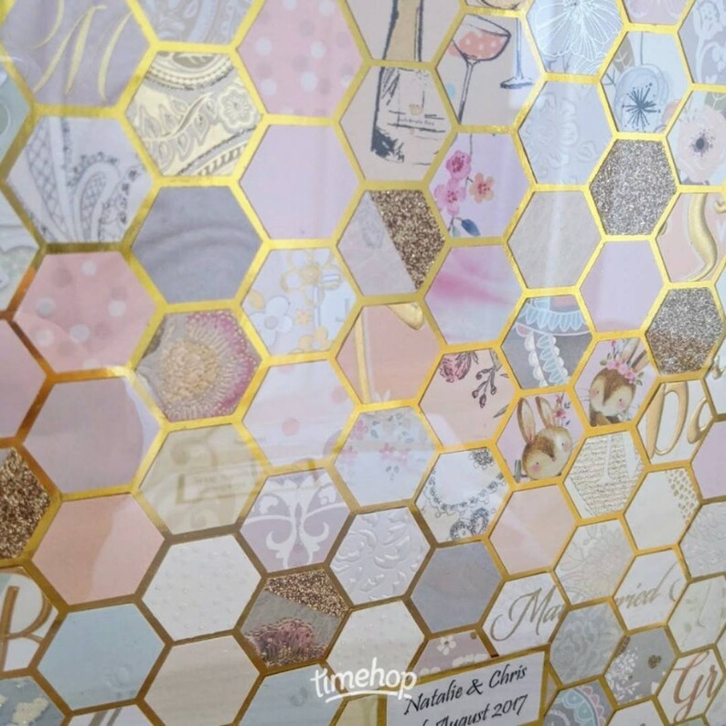 May include: A hexagonal guest book with a gold frame and a pink, white and gold patterned design. The text "Natalie & Chris Married August 2017" is printed on the bottom of the guest book.