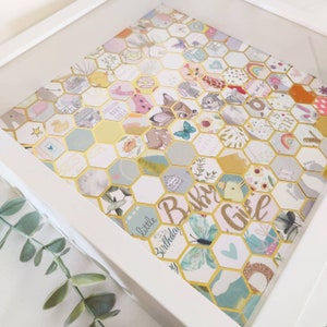 May include: A white frame containing a gold honeycomb pattern. The honeycomb pattern is filled with various images, including a deer, butterflies, and flowers. The text "Baby Girl" is written in gold glitter on the honeycomb pattern.