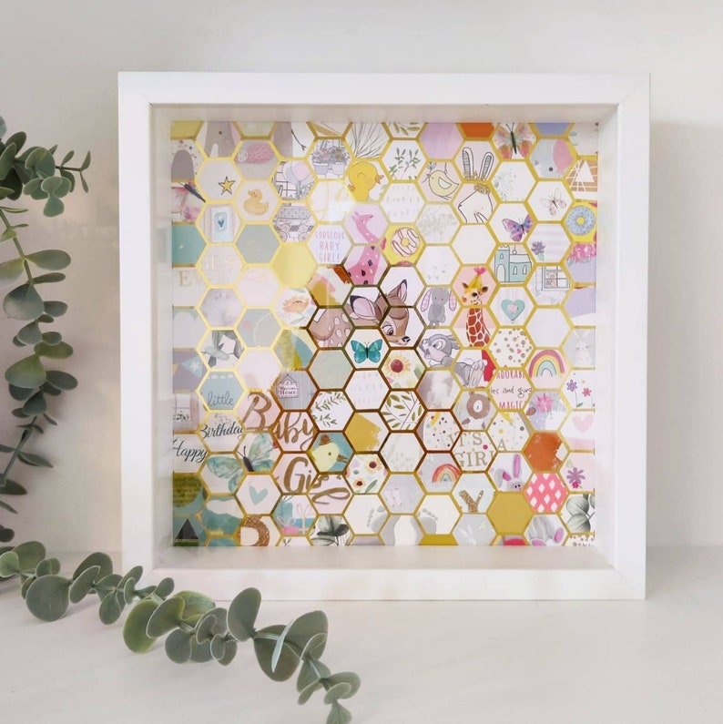 May include: A white frame with a gold honeycomb design. The honeycomb is filled with colourful paper cutouts of animals, flowers, and other baby-themed designs. The text "Baby Girl" is visible in the centre of the frame.