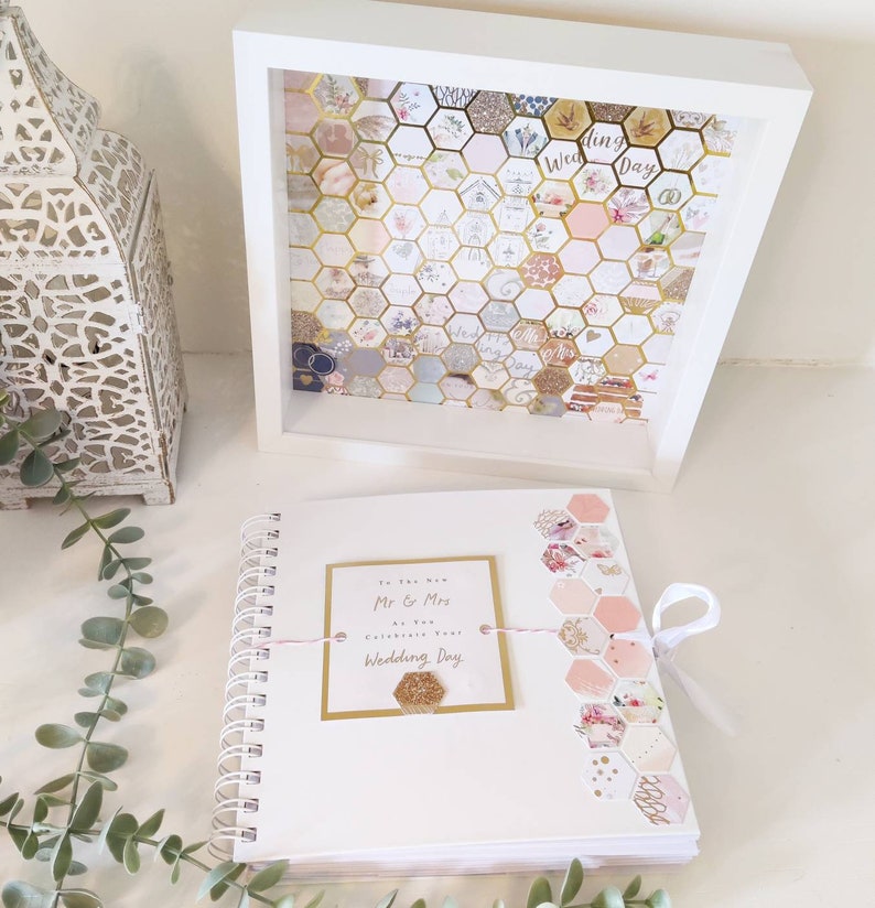 May include: A white spiral bound guest book with a pink and gold honeycomb design on the cover. The book has a tag that says "To the New Mr & Mrs A New Chapter Celebrate Your Wedding Day".