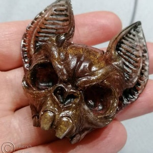 Peut inclure: Un pendentif en forme de crâne de chauve-souris brun et or avec un cordon noir. Le pendentif est en résine et présente un design détaillé.