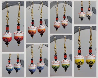 Może przedstawiać: Kolczyki wiszące z ceramicznymi koralikami w kształcie kota maneki-neko w różnych kolorach, w tym czerwonym, żółtym, niebieskim i zielonym. Każdy kolczyk ma złote haczyki, czarne i czerwone fasetowane koraliki kryształowe oraz mały czarny koralik.