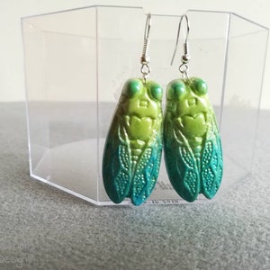 Puede incluir: Un par de pendientes de cigarra verde y azul. Los pendientes están hechos de arcilla polimérica y tienen un diseño de cigarra realista. Las cigarras son verdes con alas azules y tienen ganchos plateados.
