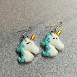 K&ouml;nnte beinhalten: Ein Paar wei&szlig;e Einhorn-Ohrringe mit goldenen H&ouml;rnern und t&uuml;rkisblauen M&auml;hnen. Die Ohrringe sind aus Polymer Clay gefertigt und haben silberne Haken.