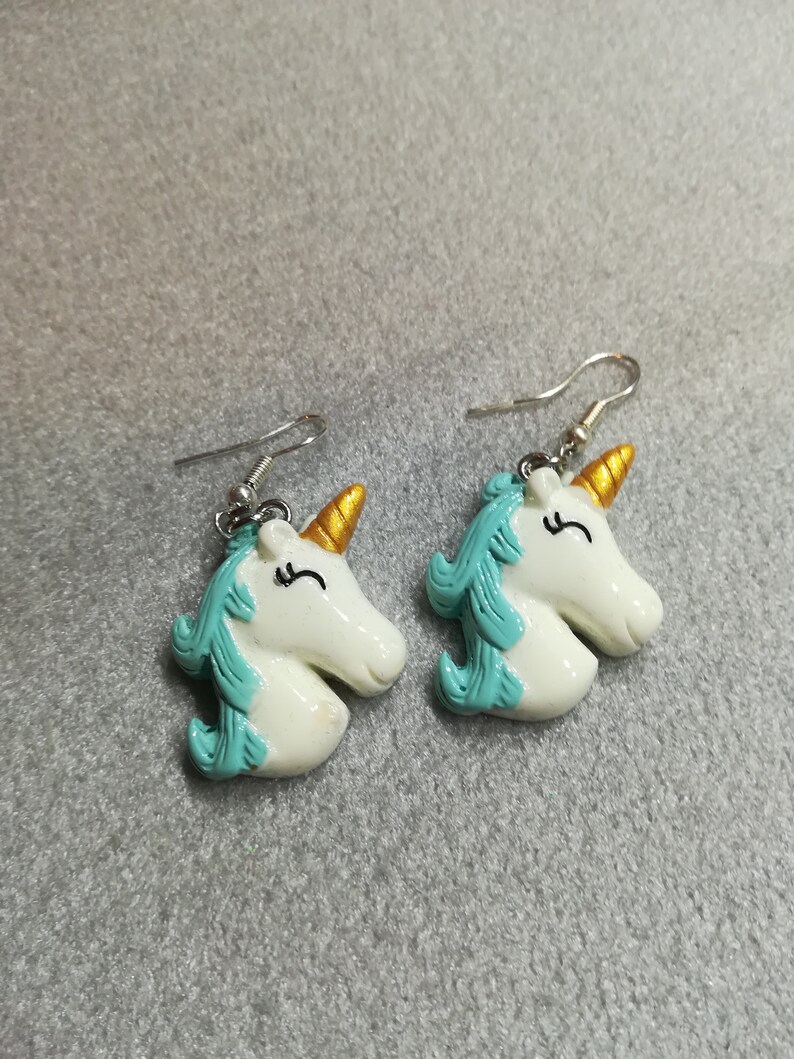 K&ouml;nnte beinhalten: Ein Paar wei&szlig;e Einhorn-Ohrringe mit goldenen H&ouml;rnern und t&uuml;rkisblauen M&auml;hnen. Die Ohrringe sind aus Polymer Clay gefertigt und haben silberne Haken.