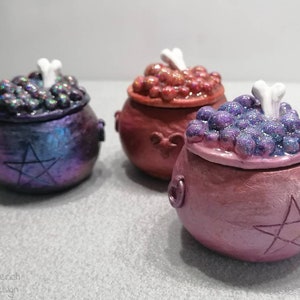 Könnte beinhalten: Drei kleine, keramikförmige Kessel mit Deckeln. Jede Box ist mit einem Pentagramm verziert und mit schimmerndem Glitzer gefüllt. Die Deckel sind mit einem weißen knochenförmigen Griff versehen.