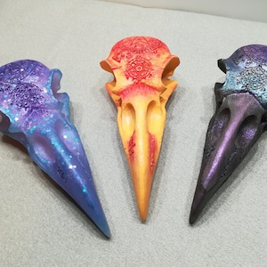 May include: Three decorative bird skulls, each painted in a different color gradient. The first skull is painted in a purple and blue gradient, the second skull is painted in a red and yellow gradient, and the third skull is painted in a dark blue and purple gradient. Each skull has intricate details and a glossy finish.