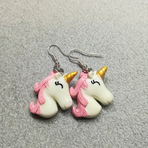 K&ouml;nnte beinhalten: Ein Paar silberne Ohrringe in Form von Einh&ouml;rnern mit rosa und goldenen Details. Die Einhornk&ouml;pfe sind wei&szlig; mit rosa M&auml;hnen und goldenen H&ouml;rnern.