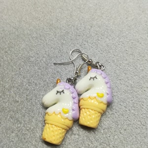 K&ouml;nnte beinhalten: Ein Paar silberne Einhorn-Eiswaffel-Ohrringe. Das Einhorn ist wei&szlig; mit lila und gelben Akzenten. Die Waffel ist gelb und wei&szlig;.