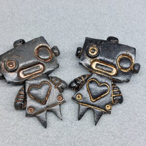 Puede incluir: Dos figuritas de robots negros con detalles dorados y en forma de corazón. Los robots están de pie sobre una superficie gris.