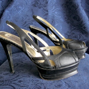 May include: A pair of black platform peep-toe slingback heels. The heels have a high stiletto heel and a platform sole. The upper features cut-out detailing and a metallic insole. The brand name "bebe" is visible on the insole.