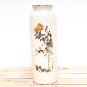 May include: A white ceramic vase with a gold rim and a floral design featuring birds and a tree. The vase has a textured surface and a delicate, hand-painted design.