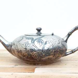May include: A silver teapot with an ornate design of grapes and leaves. The teapot has a round body and a curved spout.