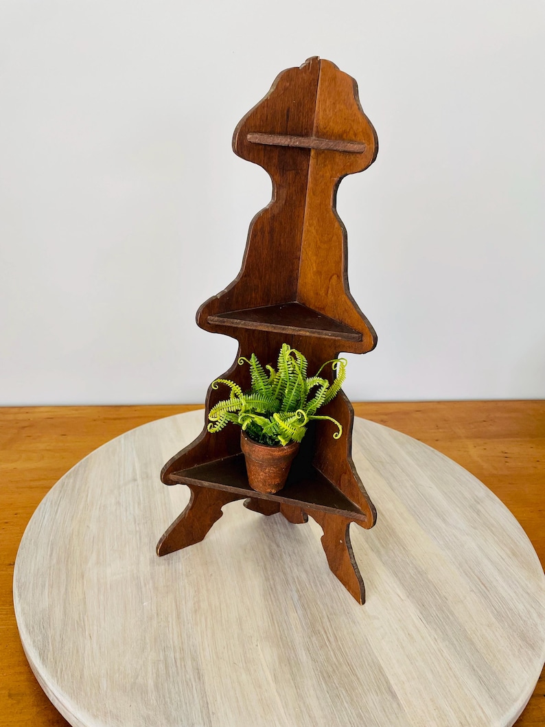 K&ouml;nnte beinhalten: Dunkelbraunes Eckregal aus Holz mit drei Ebenen und einem dekorativen Oberteil. Eine kleine Topfpflanze mit gr&uuml;nem Laub steht auf dem mittleren Regal. Das Regal steht auf einer hellen Holzoberfl&auml;che.
