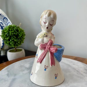 May include: A vintage ceramic figurine of a girl holding a small bird in her hands. She is wearing a white dress with a pink bow and blue and white floral accents. The figurine has a blue and white basket on the side, which can be used as a planter.