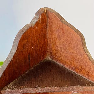 K&ouml;nnte beinhalten: Nahaufnahme eines dekorativen Holzobjekts mit dreieckiger Form. Das Holz hat eine rotbraune Farbe mit sichtbarer Maserung. Die Kanten sind leicht abgerundet und haben einen helleren Ton.