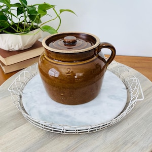 May include: A brown ceramic crock with a lid and handle. The crock is round with a slightly tapered shape and a number "2" is visible on the side. It sits on a white marble tray inside a wire basket.