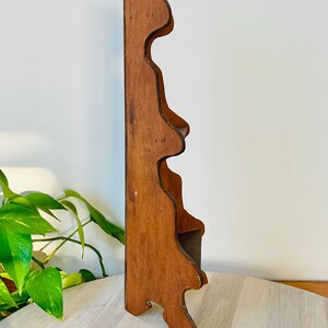 K&ouml;nnte beinhalten: Ein h&ouml;lzernes Wandregal mit warmbraunem Finish. Das Regal hat eine dekorative, wellenf&ouml;rmige Kante und eine kleine Ablagefl&auml;che. Das Design ist vintage und eignet sich zum Pr&auml;sentieren kleiner Gegenst&auml;nde oder Dekoration. Ca. 30 cm hoch.