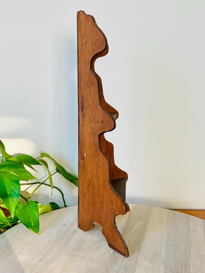 K&ouml;nnte beinhalten: Ein h&ouml;lzernes Wandregal mit warmbraunem Finish. Das Regal hat eine dekorative, wellenf&ouml;rmige Kante und eine kleine Ablagefl&auml;che. Das Design ist vintage und eignet sich zum Pr&auml;sentieren kleiner Gegenst&auml;nde oder Dekoration. Ca. 30 cm hoch.