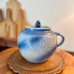 May include: A blue and white ceramic jar with a lid and handle. The jar has embossed text and sits on a wooden tray. The jar is round with a small knob on the lid.