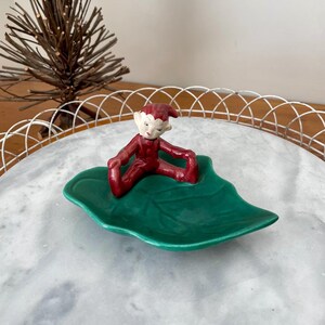 May include: A ceramic figurine of a seated elf in a red outfit with a matching hat. The elf is resting on a green leaf-shaped dish. The dish is placed on a white marble tray.