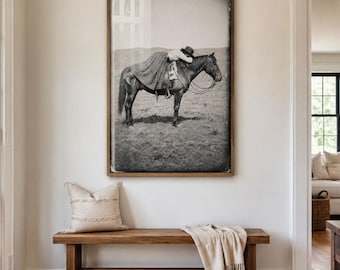 May include: A large rectangular framed black and white photograph depicting a person wearing a hat and long skirt resting on a horse in a field. The artwork is mounted on a plain wall above a wooden bench with a beige pillow and a draped throw blanket.