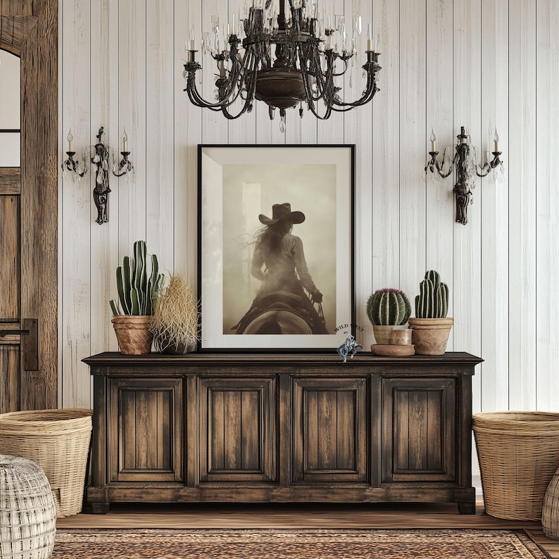 May include: A rectangular framed wall art print featuring a sepia-toned photograph of a person in a cowboy hat riding a horse. The artwork is centered on a dark brown wooden sideboard with four cabinet doors, flanked by potted cacti in clay pots.