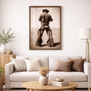May include: A framed sepia-toned wall art print depicting a person from behind, wearing a wide-brimmed hat, a jacket, and leather chaps while standing in a vast, arid desert landscape. The artwork is displayed in a dark wooden frame.