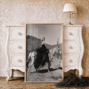 May include: A framed black and white photograph featuring a person with blonde hair wearing a dark strapless dress, riding a spotted horse in a desert landscape with cacti and mountains in the background. The print is displayed in a light wood frame.