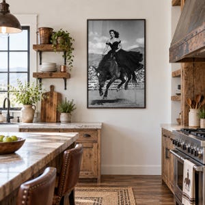 May include: A framed black and white wall art print depicting a person in a long dark dress riding a horse mid-jump. The artwork is displayed on a light-colored wall in a kitchen featuring wooden cabinets, open shelving, and a stainless steel stove.
