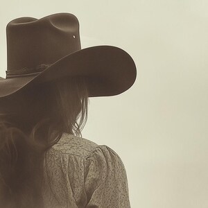 May include: A dark brown cowboy hat with a wide, curved brim and a creased crown, worn over long hair. The hat features a thin, dark band around the base of the crown. The overall tone of the image is sepia, highlighting the texture of the hat.