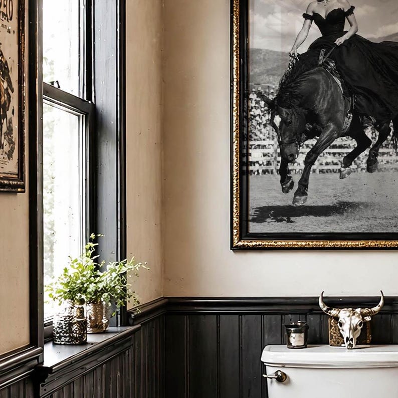 May include: A framed black and white photograph of a person in a dark gown riding a horse, displayed on a wall above a toilet. The room features dark wood wainscoting, a window with a small green plant, and a decorative animal skull on the tank.