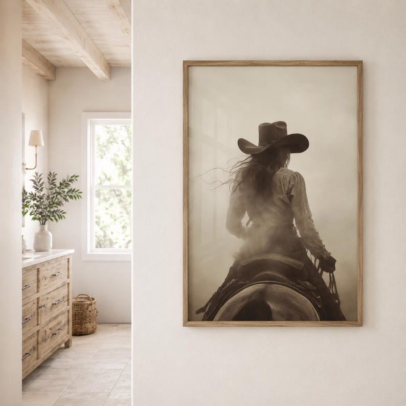 May include: A framed wall art print featuring a sepia-toned photograph of a person wearing a wide-brimmed cowboy hat, seen from behind while riding a horse through a dusty, hazy landscape. The artwork is displayed in a light wood frame.
