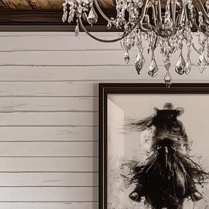May include: A framed black and white wall art print depicting a person wearing a cowboy hat riding a horse, displayed on a white horizontal shiplap wall beneath a crystal chandelier with multiple hanging glass teardrop prisms and metal arms.