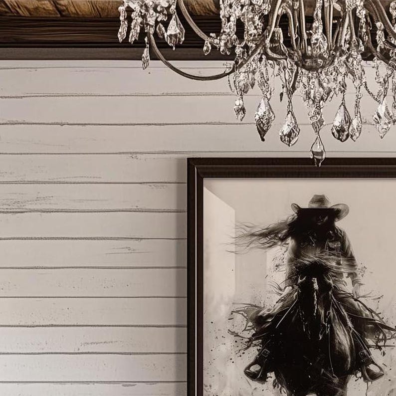 May include: A framed black and white wall art print depicting a person wearing a cowboy hat riding a horse, displayed on a white horizontal shiplap wall beneath a crystal chandelier with multiple hanging glass teardrop prisms and metal arms.