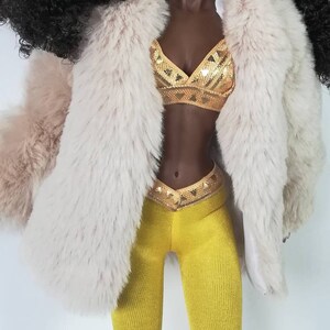 Puede incluir: Una mu&ntilde;eca con el pelo largo y negro rizado que lleva un abrigo de piel sint&eacute;tica blanca, un top de lentejuelas doradas y leggings amarillos.
