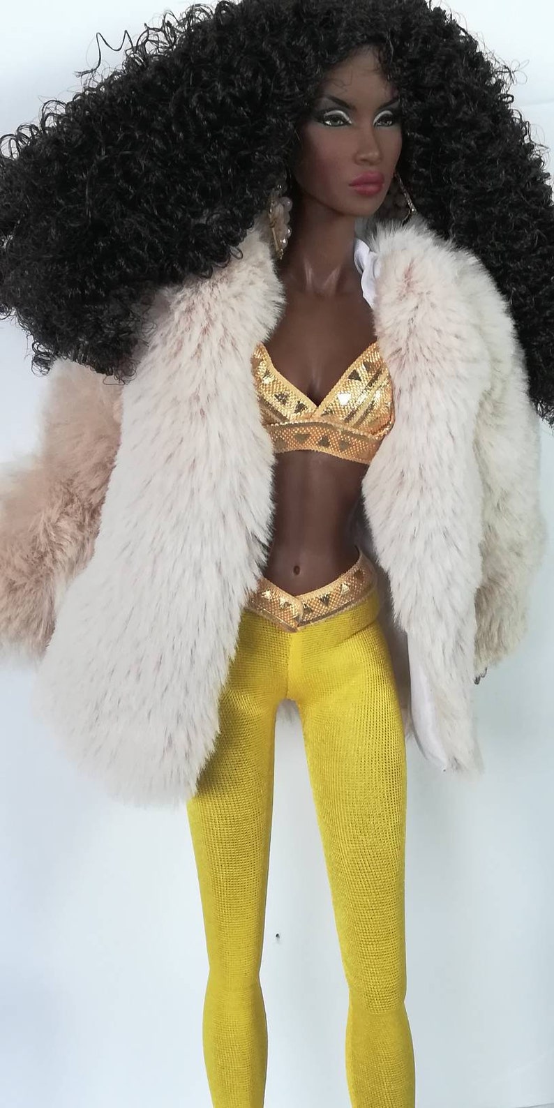 Puede incluir: Una mu&ntilde;eca con el pelo largo y negro rizado que lleva un abrigo de piel sint&eacute;tica blanca, un top de lentejuelas doradas y leggings amarillos.