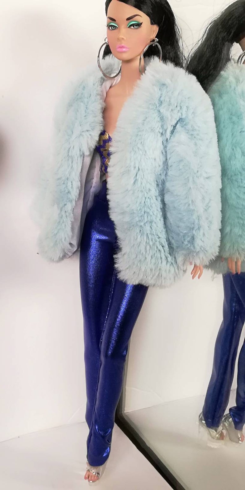 May include: A doll wearing a light blue faux fur jacket and blue metallic trousers. The doll has long black hair and is wearing silver hoop earrings.